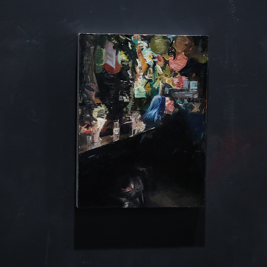 A square dark painting or print depicting a crowded street scene with colorful figures and objects, mounted against a very dark wall with a soft shadow.