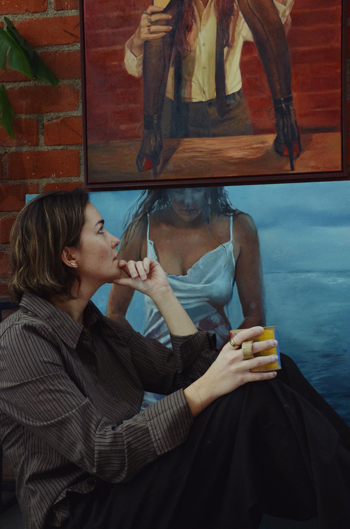 Sapphic artist Halie Torris sitting beside her original figurative oil painting in her Los Angeles art studio.